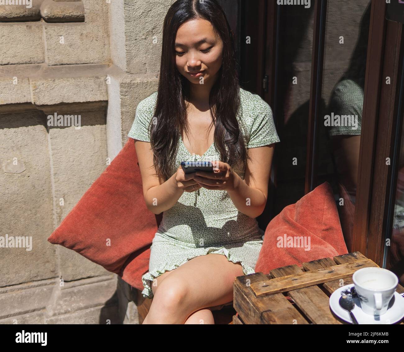 Attraktive asiatische weibliche Surfen Handy während sitzen am Tisch mit leeren Tasse Kaffee auf der Terrasse der Cafeteria im Sommer Stockfoto