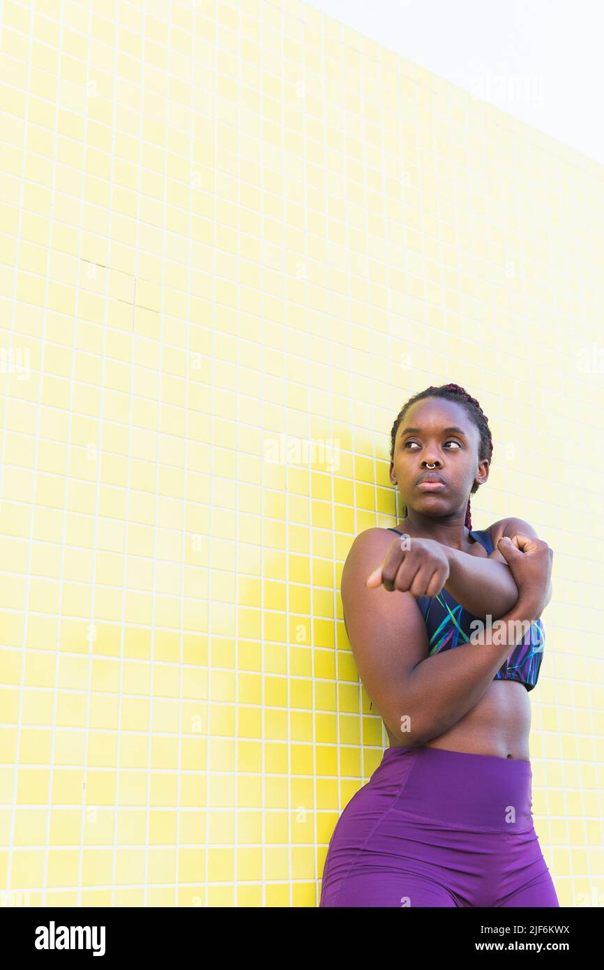Passen Sie die schwarze Sportlerin in die Sportswear und machen Sie Stretching-Übungen, während Sie sich vor dem Training an der gelb gefliesten Wand aufwärmen Stockfoto