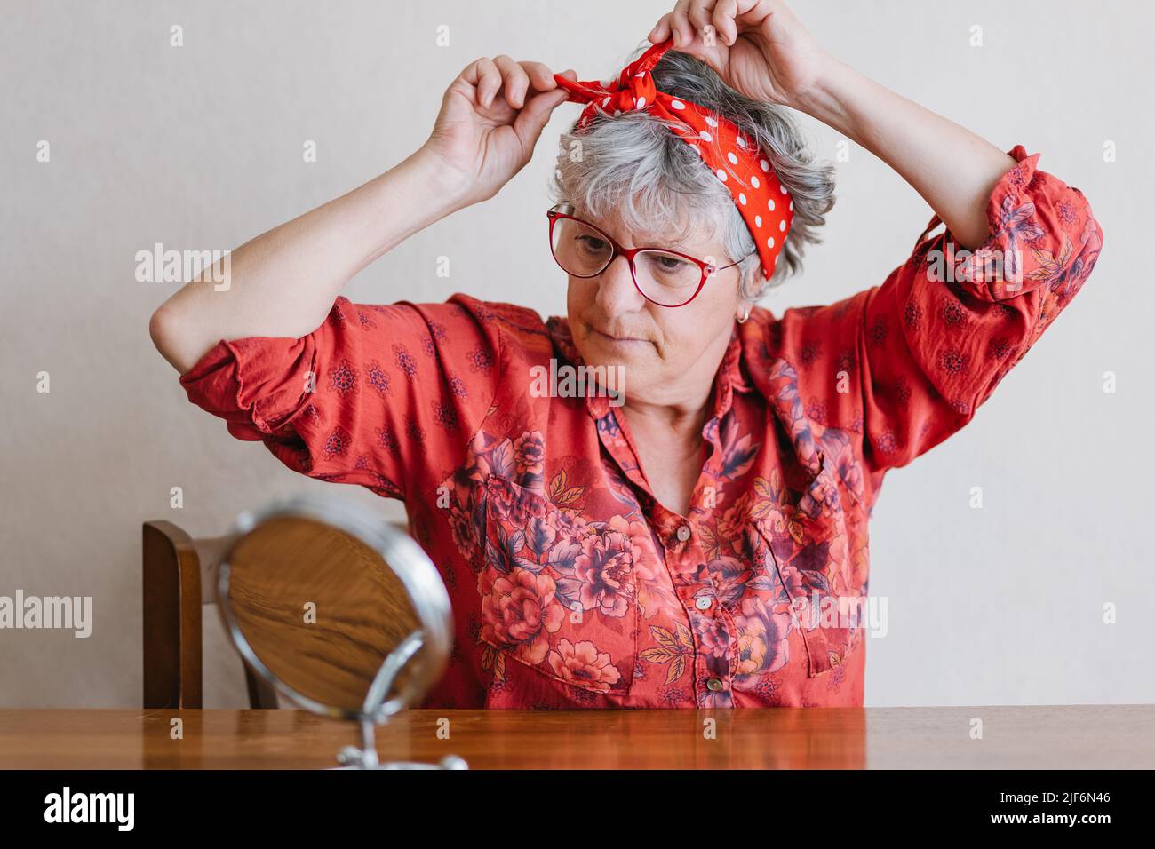 Stilvolle ältere Frau mit grauem Haar in eleganter Bluse und Brillen, die das Stirnband anpassen und in runden Kosmetikspiegel schauen, der zu Hause am Tisch sitzt Stockfoto