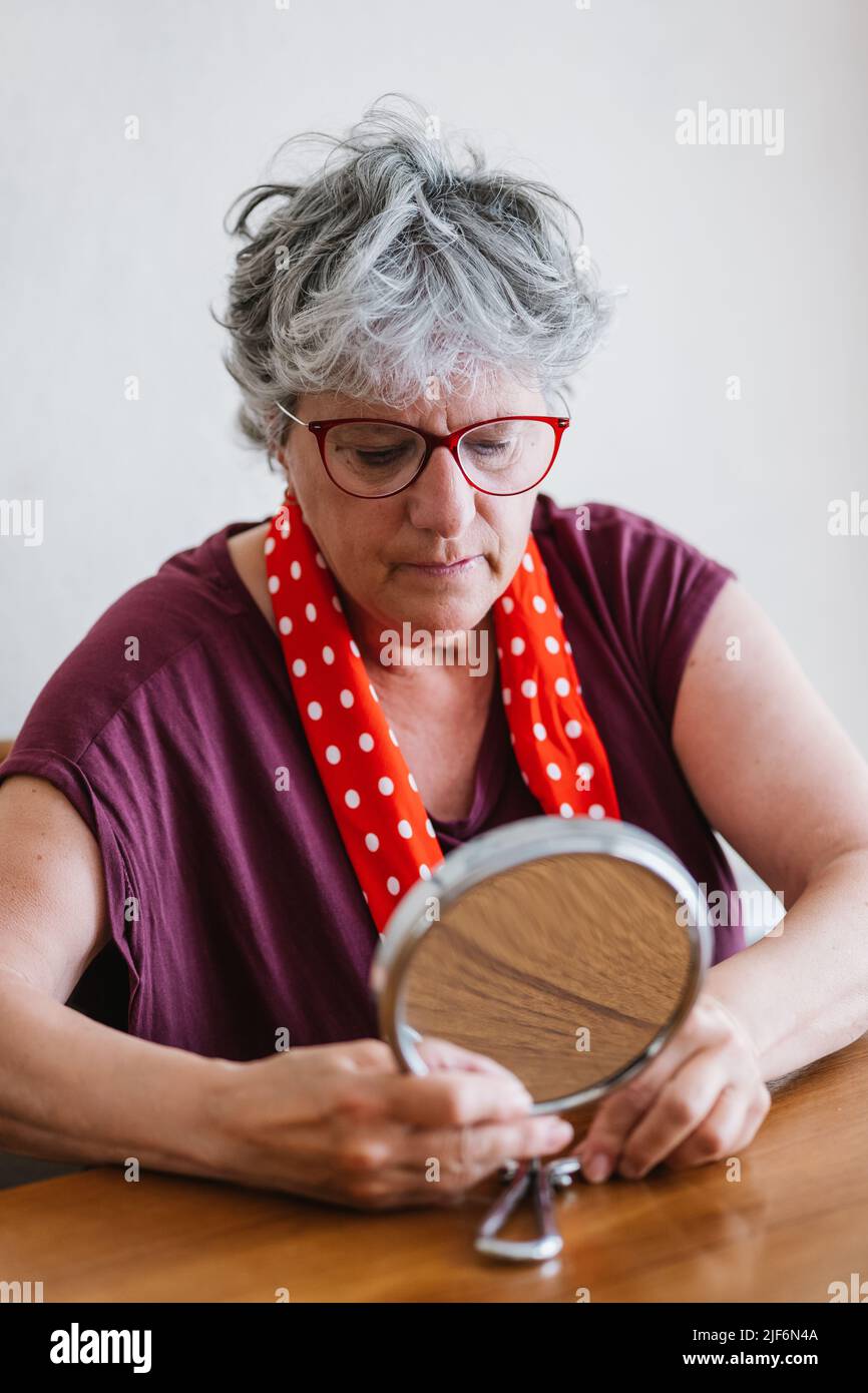 Konzentrierte ältere Frau mit grauen Haaren in legerem Outfit und Brille, die am Holztisch sitzt und in einem runden Kosmetikspiegel zu Hause schaut Stockfoto