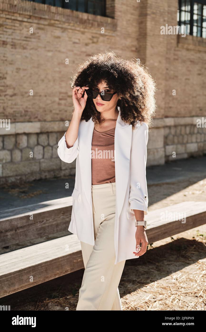 Trendige hispanische Unternehmerin in weißem Anzug mit lockigen Haaren, die Sonnenbrille auspasst und beim Spaziergang in der Nähe eines schäbigen Gebäudes auf Cit auf die Kamera schaut Stockfoto