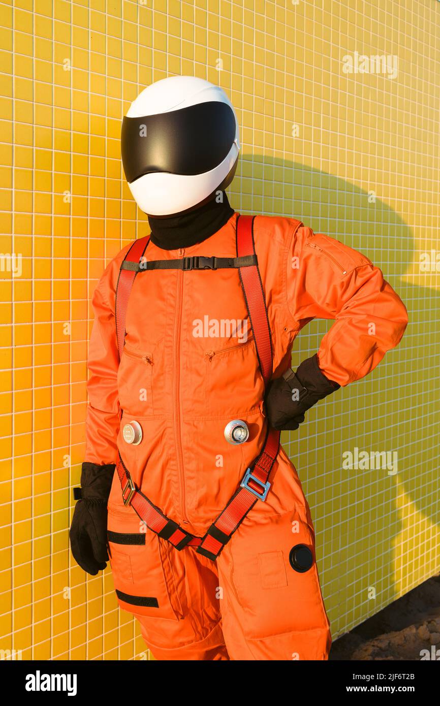 Selbstsicherer Astronaut in Raumanzug und Helm, der die Hände an der Taille hält und auf Sand an der gefliesten gelben Wand am Strand steht Stockfoto