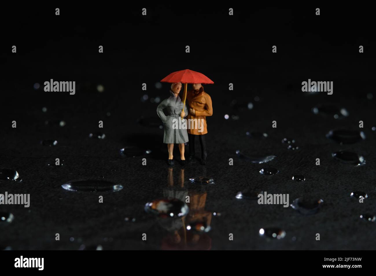 Miniatur Menschen Spielzeug Figur Fotografie. Ein Paar, das einen Sonnenschirm benutzt, läuft auf dem Boden voller Tropfen. Dunkler wolkig Himmel Hintergrund. Bildfoto Stockfoto