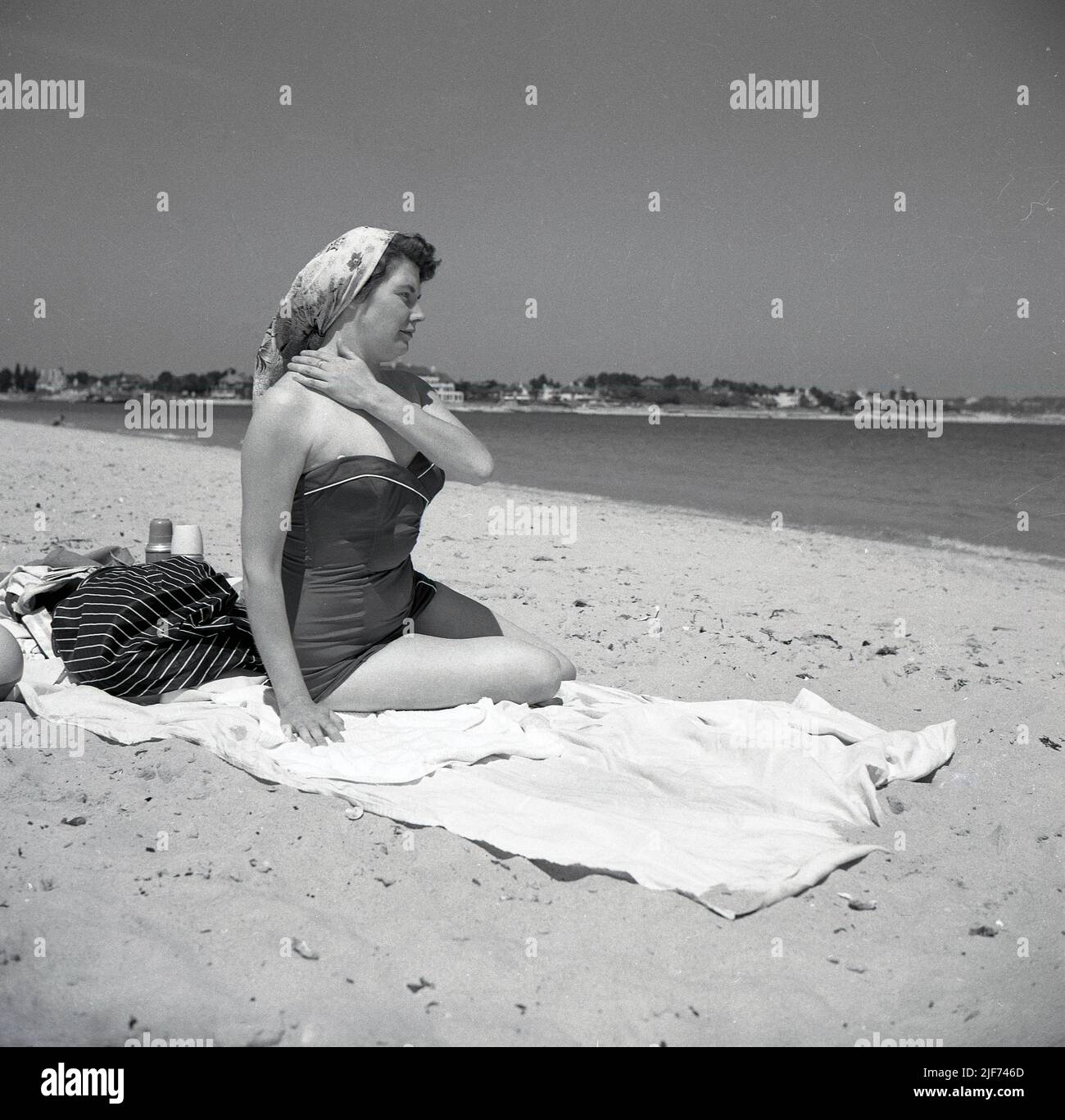 1950s, historisch, eine Frau in einem Badeanzug der Zeit, mit einem Kopftuch, sitzt auf einem Handtuch auf einem Sandstrand, England. VEREINIGTES KÖNIGREICH. Stockfoto