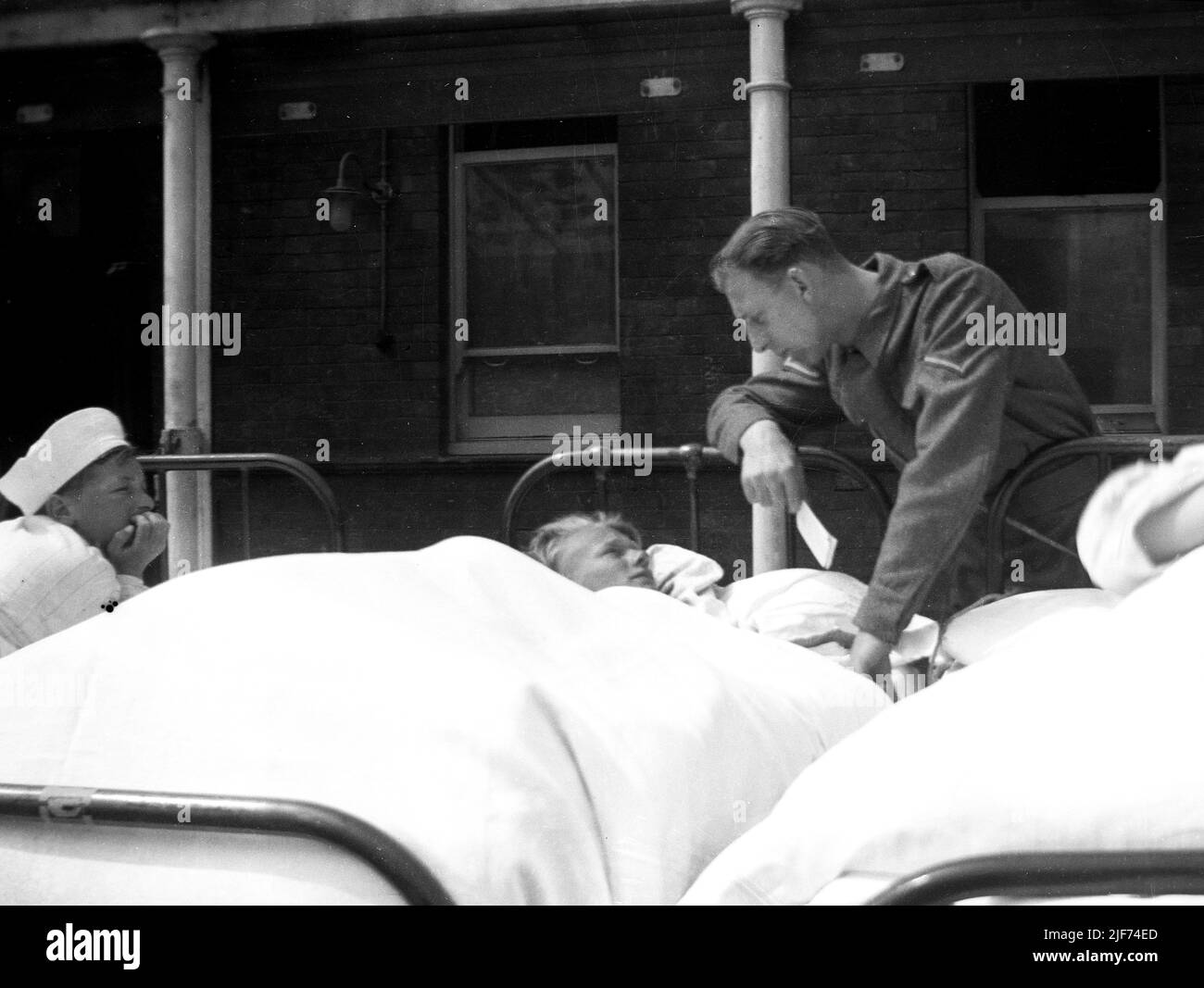 1941, historisch, WW2 und ein uniformierte Soldat der britischen Armee, ein Lance Corporal, im Gespräch mit einem jungen Jungen, der in einem Metallkrankenhausbett vor dem Queen Mary's Hospital for Children, Carshalton, England, Großbritannien, liegt. 1908 als Southern Hospital erbaut, wurde es 1909 zu einem Kinderkrankenhaus und nach einem Besuch von Queen Mary im Jahr 1915 umbenannt. Es war das am stärksten bombardierte Krankenhaus im Großraum London, mit dem ersten Angriff im Jahr 1940. Die neuen Bedrohungen durch die VI- und V2-Raketen fanden im Juli 1944 eine totale Evakuierung statt, bei der Kinder an andere Orte in England und Wales gebracht wurden. Stockfoto