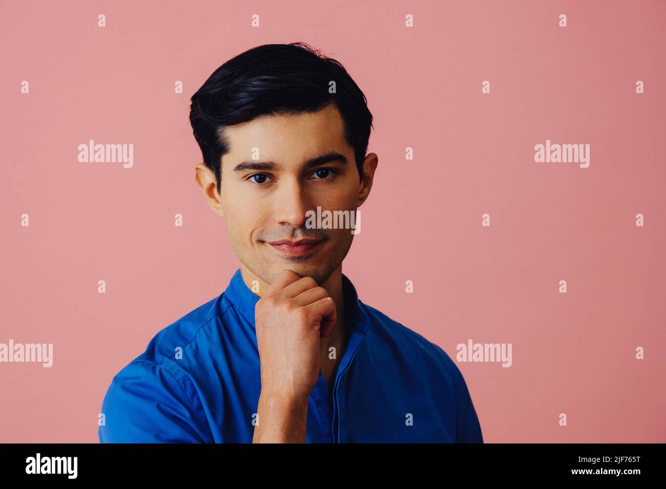 Headshot nachdenklicher, hübscher, junger, erwachsener Latino-Mann mit Hand am Kinn, schwarzen Haaren und blauem Hemd auf rosa Hintergrund, der auf das Kamerastudio schaut Stockfoto