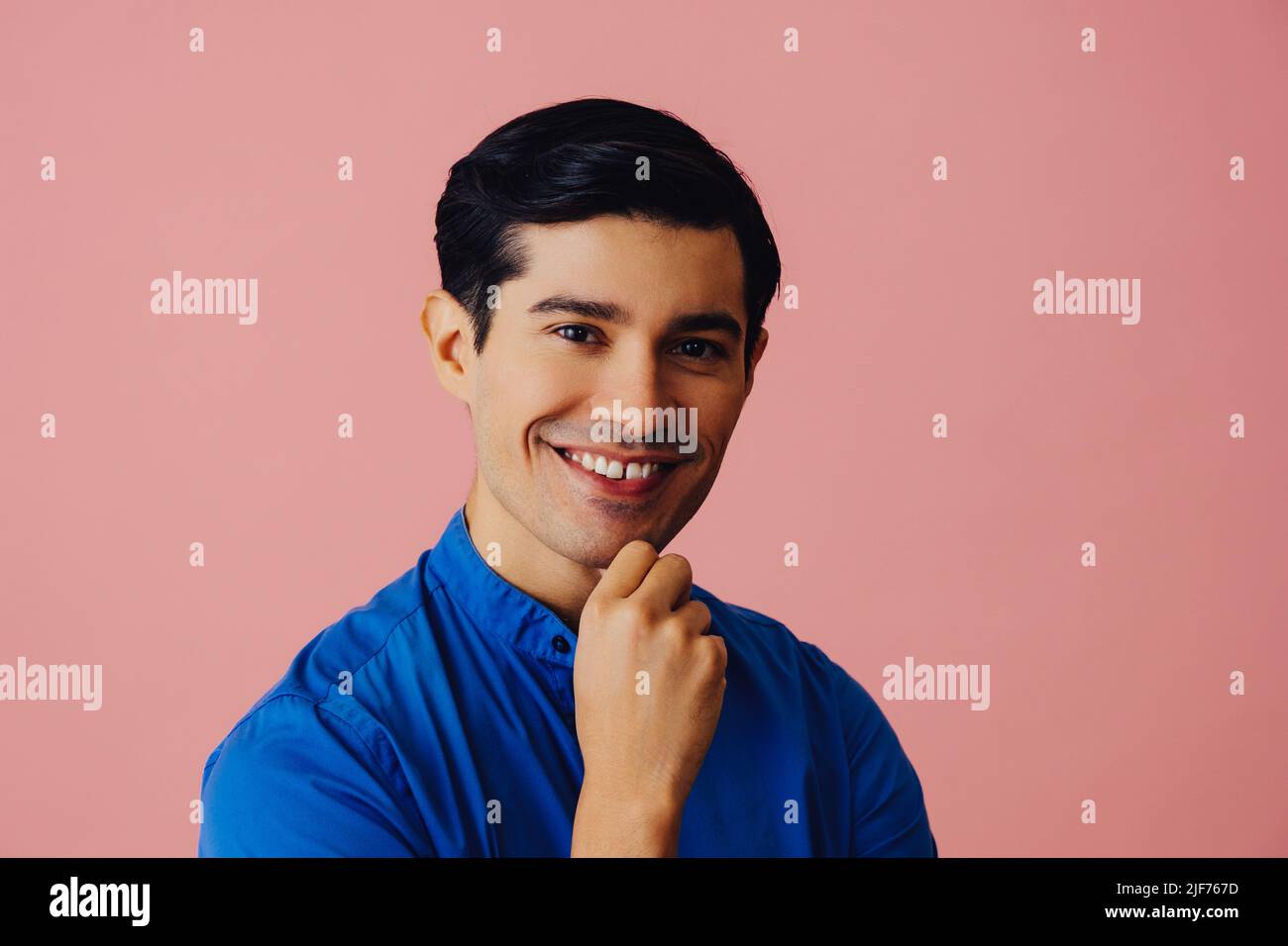 Kopfschuss lächelnd schöner junger Erwachsener Latino Mann mit Hand auf dem Kinn schwarzen Haaren und blauem Hemd über rosa Hintergrund Blick auf Kamera Studio aufgenommen Stockfoto