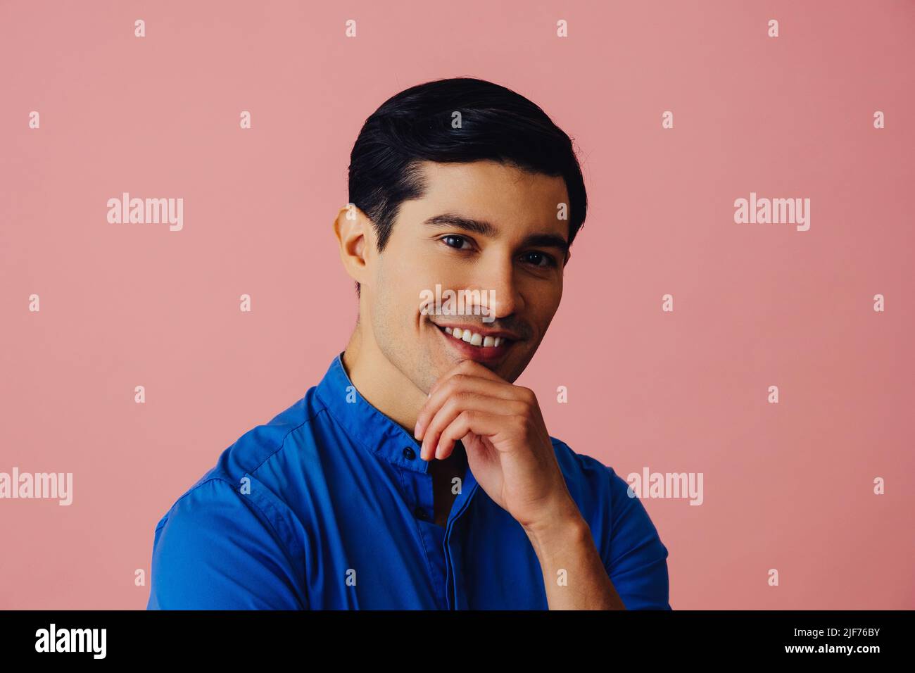 Kopfschuss lächelnd schöner junger Erwachsener Latino Mann mit Hand auf dem Kinn schwarzen Haaren und blauem Hemd über rosa Hintergrund Blick auf Kamera Studio aufgenommen Stockfoto