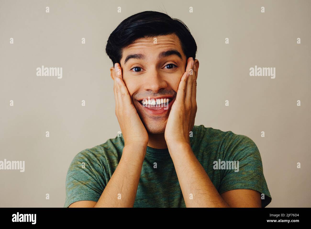 Headshot hispanic Latino Mann lächelnde Hände auf Wange schwarzes Haar Hand bedeckt Mund schönes junges Erwachsener grünes T-Shirt über grauem Hintergrund Blick auf Kamera Studio aufgenommen Stockfoto