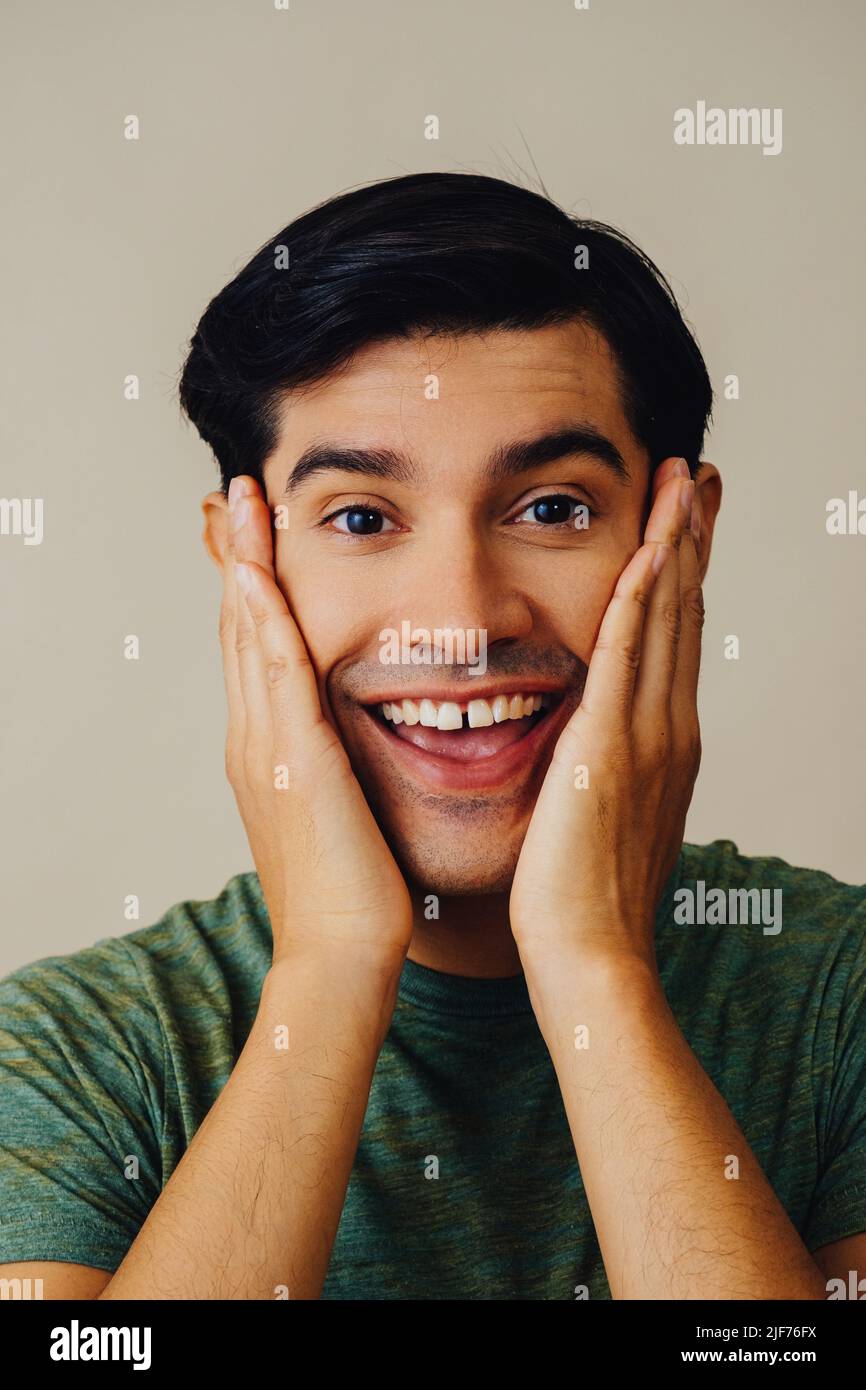 Headshot hispanic Latino Mann lächelnde Hände auf Wange schwarzes Haar Hand bedeckt Mund schönes junges Erwachsener grünes T-Shirt über grauem Hintergrund Blick auf Kamera Studio aufgenommen Stockfoto