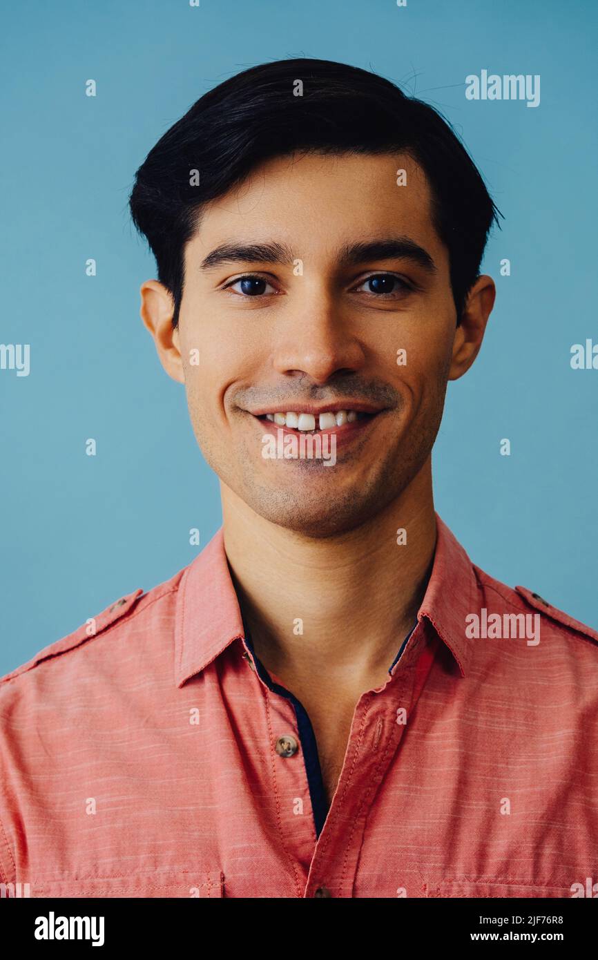 Headshot hispanic Latino Mann schwarze Haare lächelnd schöner junger Erwachsener trägt rosa Hemd über blauem Hintergrund Blick auf Kamera Studio aufgenommen Stockfoto