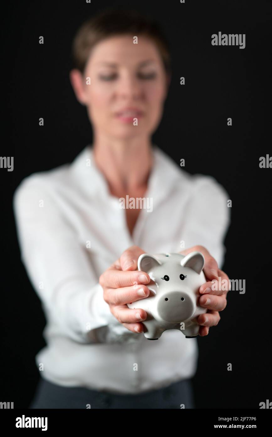 Weißes Schweinebank aus Porzellan, das von einer Frau in Geschäftskleidung, weißem Hemd und grauem Rock auf schwarzem Hintergrund gehalten wird. Konzept für Geld, Finanzen, Einsparungen Stockfoto