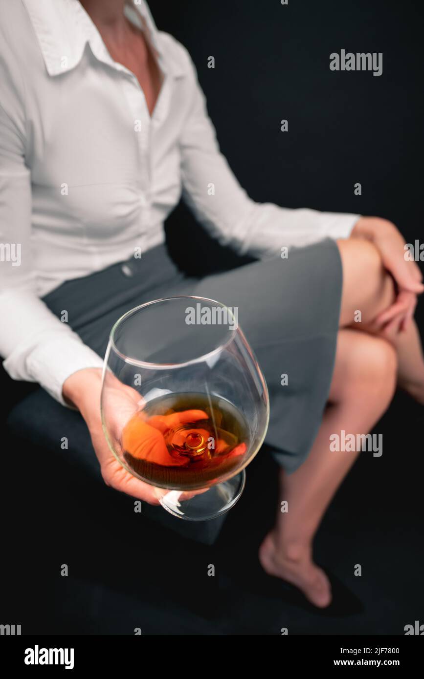 Trinkglas gefüllt mit Alkohol, das von einer Frau in Geschäftskleidung mit gekreuzten Beinen gehalten wird. Frauenbild verschwommen. Konzept für Depressionen, traurig Stockfoto
