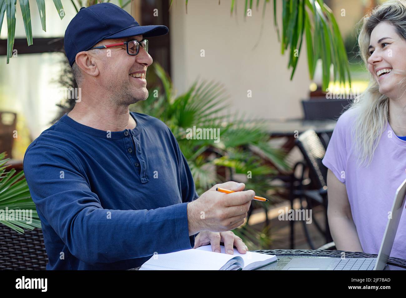 Porträt einer glücklichen Familie. Mann mittleren Alters, der mit einem Laptop im Café am Tisch sitzt, den Stift hält und mit einer jungen Frau lacht. Stockfoto