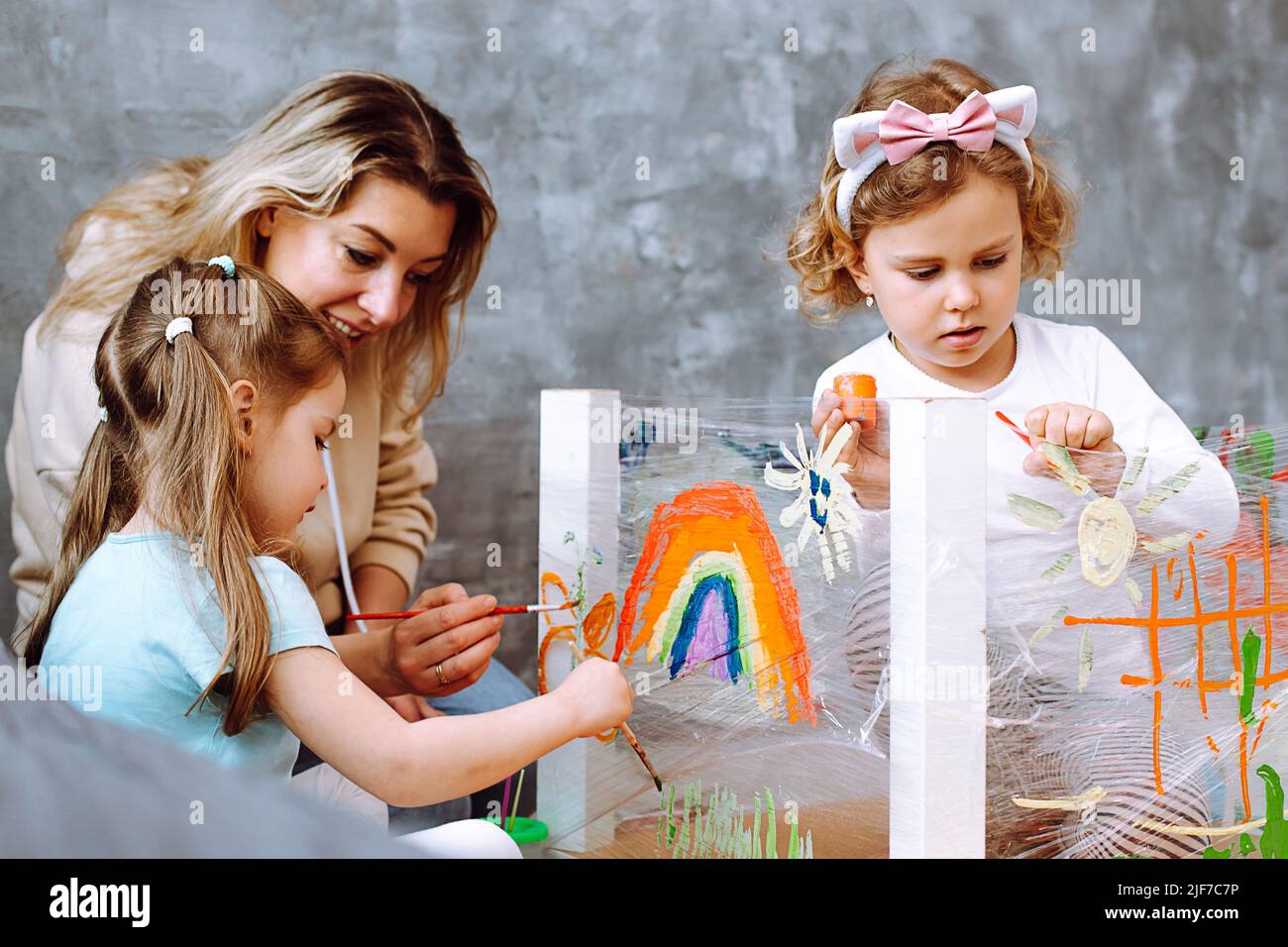 Erzieherin, die Unterricht mit Kindern im Zeichnen im Kindergarten durchführt. Mädchen sitzen und malen mit Pinsel im Spielzimmer. Kinder Stockfoto