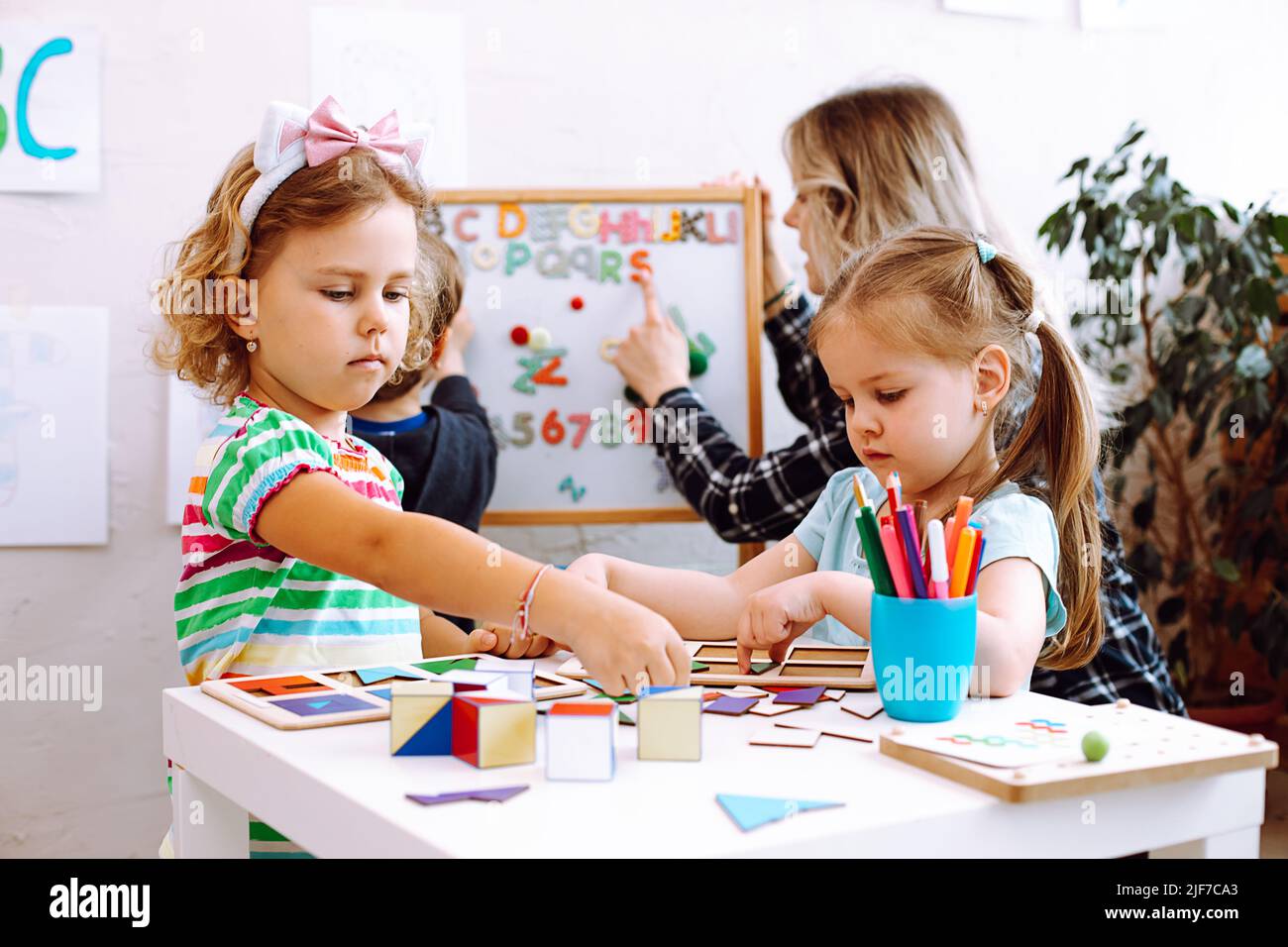 Kleine Mädchen spielen mit Würfeln und geometrischen Figuren auf dem Schreibtisch im Spielzimmer, Erzieherin mit Jungen, der Alphabet auf dem Hintergrund studiert. Interessante Lektion für Stockfoto