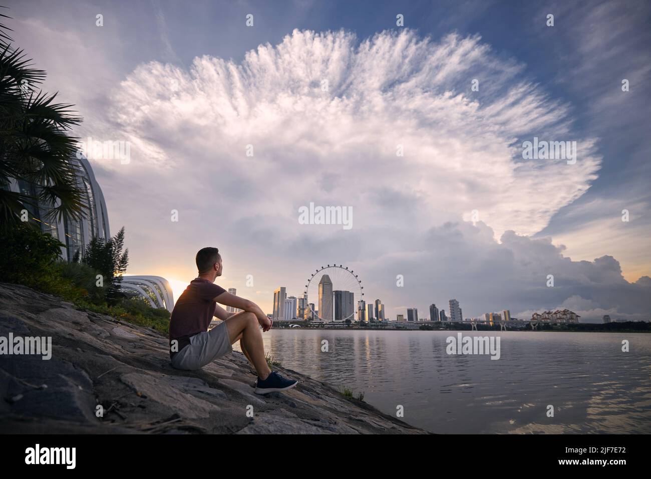 Junger Mann, der am Wasser sitzt und dramatische Gewitterwolken über der Skyline von Singapur sieht. Stockfoto