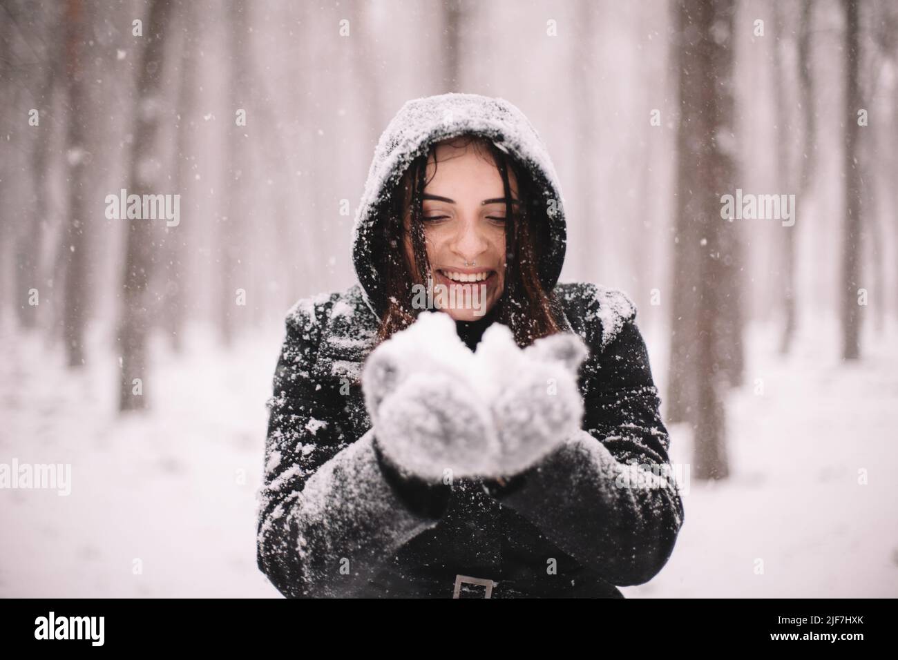 Glückliche lächelnde junge Frau, die während des Schneefalls im Winter Schnee hält Stockfoto