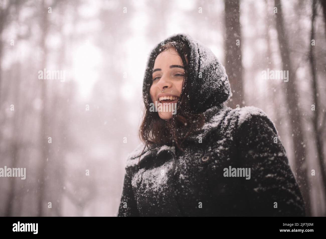 Porträt einer glücklichen jungen Frau, die während des Schneefalls im Wald steht Stockfoto