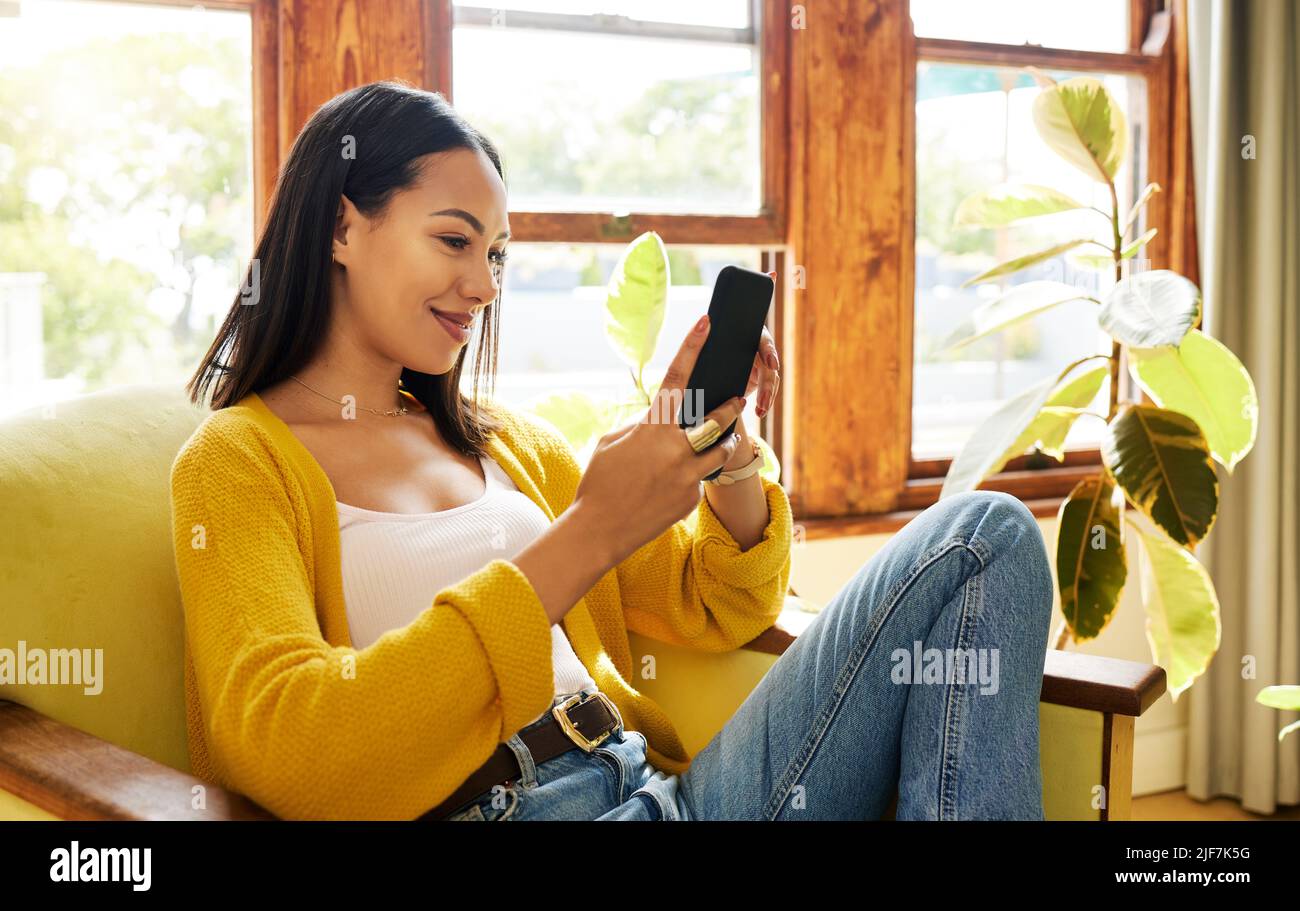 Hispanische Frau mit ihrem Smartphone, das vor einem Fenster in einem hellen Wohnzimmer sitzt. Eine junge glückliche Frau lächelt an ihrem Handy und entspannt sich auf einem Stockfoto