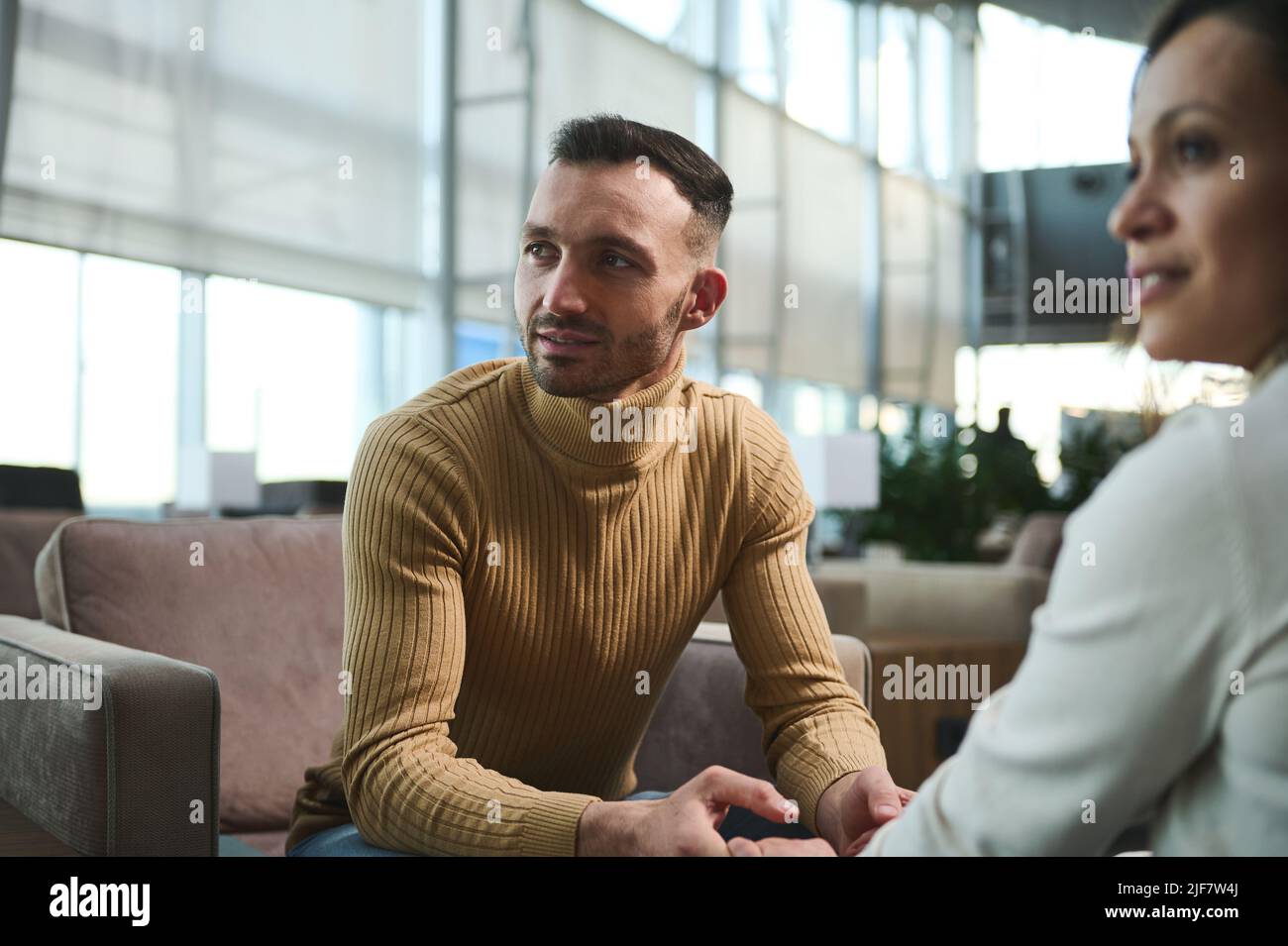 Konzentrieren Sie sich auf einen gutaussehenden arabischen Mann im Pullover, der wegschaut, die Hände seiner Freundin hält und in der VIP-Lounge des International airpo auf einem Stuhl sitzt Stockfoto
