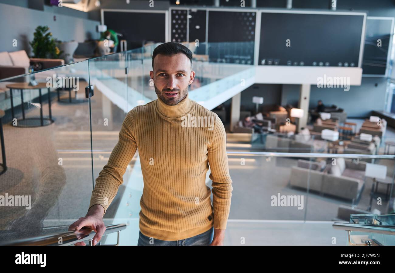 Ein gutaussehender, selbstbewusster junger Mann aus dem Nahen Osten blickt selbstbewusst in die Kamera, während er in der VIP-Lounge des internationalen Flughafens die Treppe hinaufgeht Stockfoto