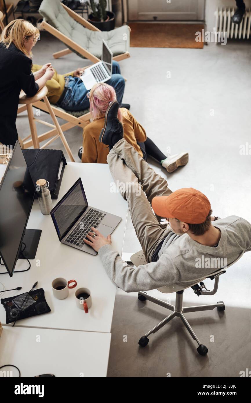 Männlicher Unternehmer mit den Füßen auf dem Schreibtisch und Laptop von Kollegen, die im Tech-Start-up-Büro diskutieren Stockfoto