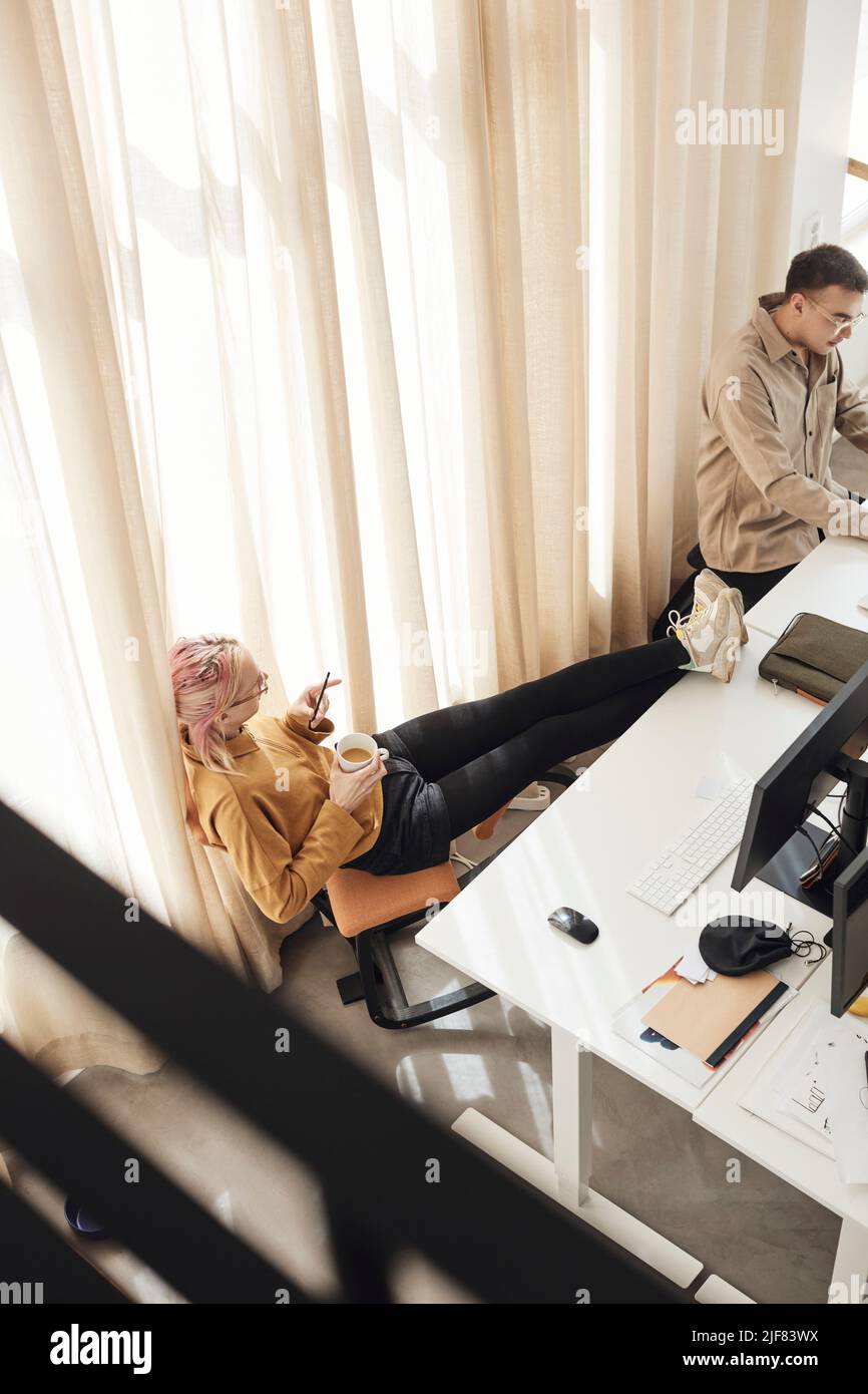 Ein männlicher Kollege im Büro hat einen Blick auf einen nicht-binären Unternehmer aus der Perspektive eines Mannes, der die Füße auf dem Schreibtisch hochgestellt hat Stockfoto