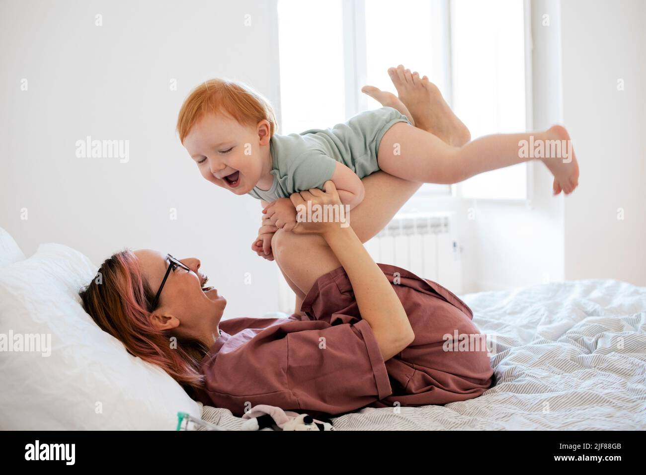 Fröhliche Mutter pflückt einen Jungen, während sie im Schlafzimmer auf dem Bett liegt Stockfoto