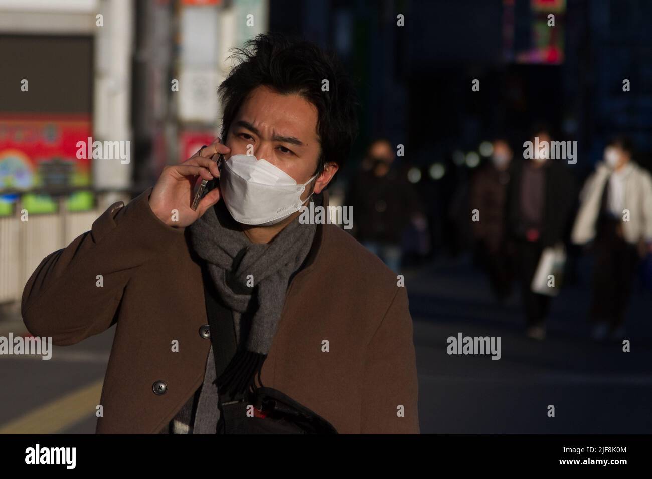 Ein Japaner, der eine OP-Maske trägt, spricht auf seinem Handy, während er in Shinjuku, Tokio, Japan, auf der Straße geht. Stockfoto