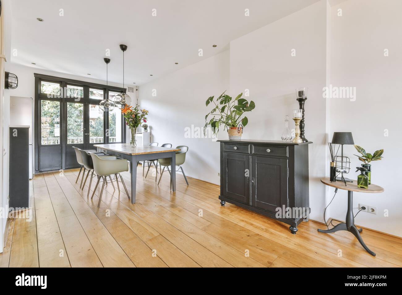 Stilvolles Wohndesign der Essecke mit Lampen, die über Holztisch und -Stühlen hängen, in einem modernen Apartment mit großen Fenstern und minimalistischem Design Stockfoto