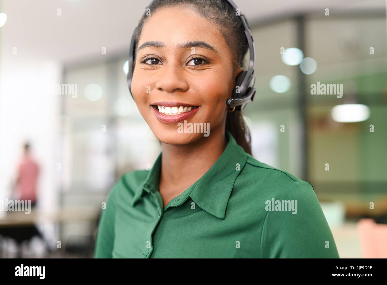 Headshot einer jungen afroamerikanischen Mitarbeiterin, die ein schnurloses Headset trägt und lächelnd auf die Kamera blickt. Fröhliche Frau in Kopfhörern mit Mikrofon, Supportmitarbeiter im Büro Stockfoto