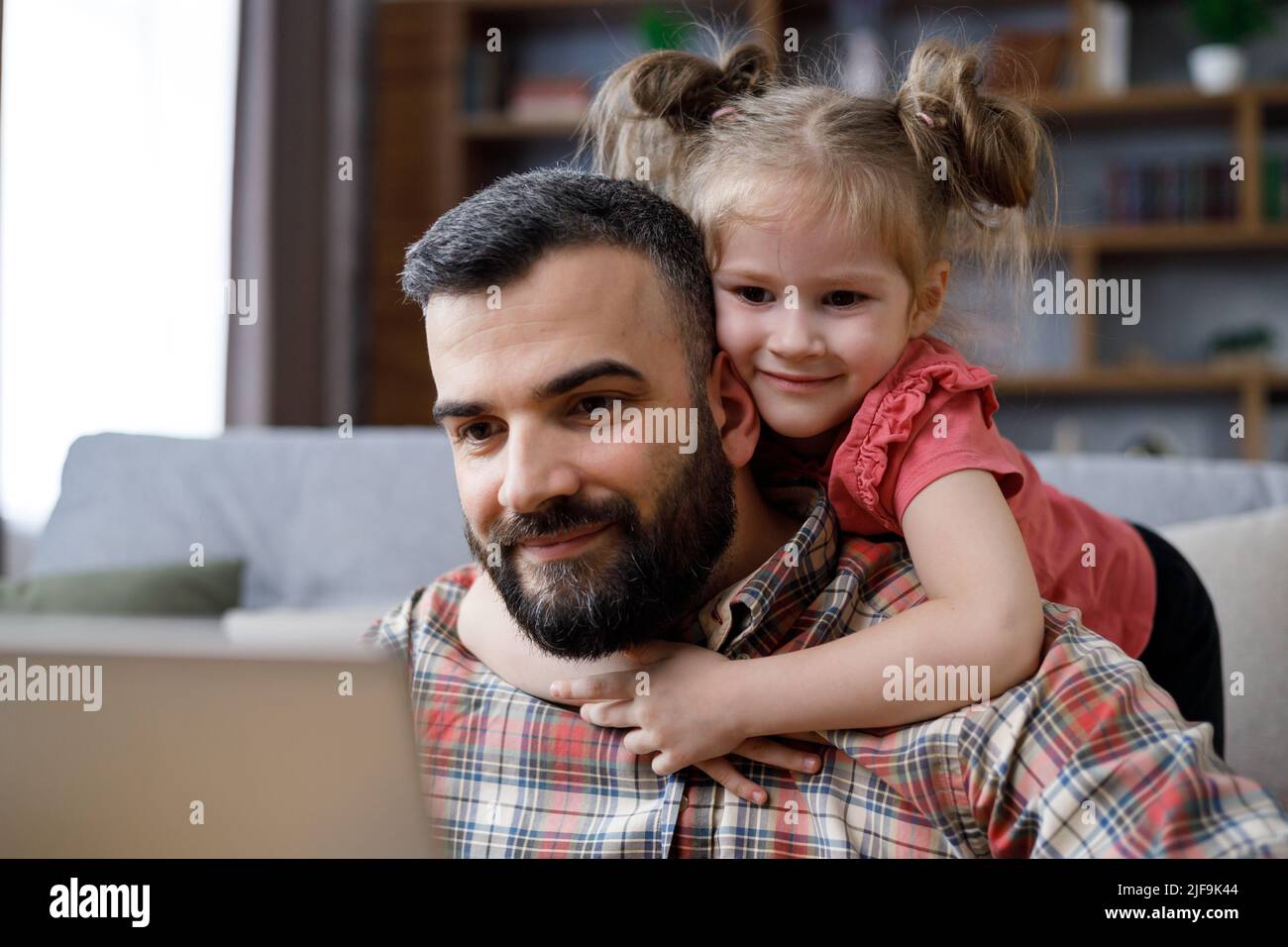 Glückliches Familienportrait. Der junge, gutaussehende Mann arbeitet von zu Hause aus am Laptop, konzentriert die Aufmerksamkeit auf den Laptop-Bildschirm, während seine kleine Tochter ihre Geliebte umarmt Stockfoto