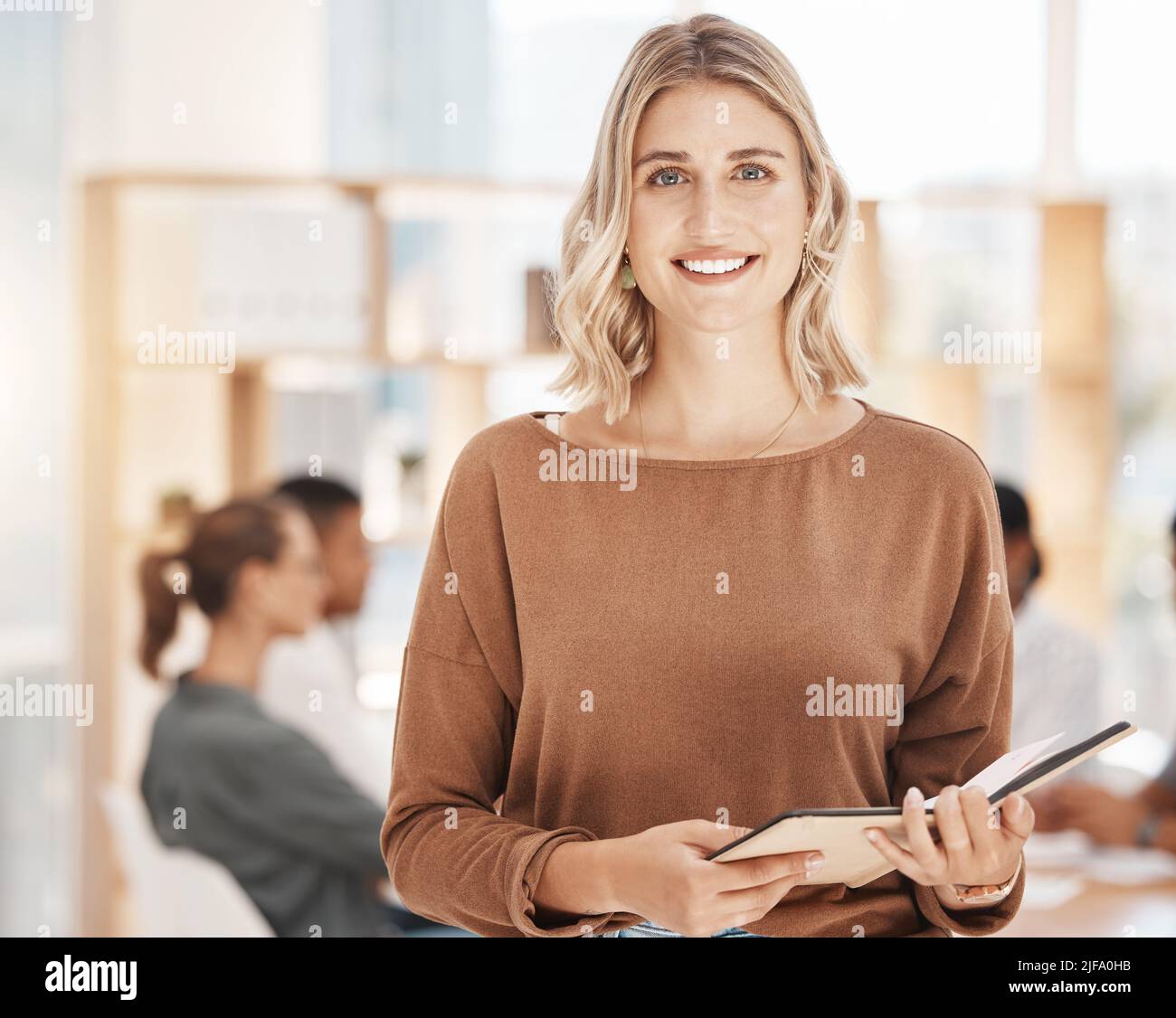 Porträt einer selbstbewussten jungen kaukasischen Geschäftsfrau, die einen Notizbuchplaner in einem Büro mit ihren Kollegen im Hintergrund hält. Ehrgeizig Stockfoto