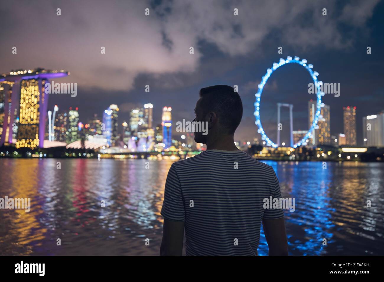 Ein hübscher Mann, der am Ufer der Bucht steht und nachts die beleuchtete Stadt anschaut. Städtische Skyline mit Bokeh von Lichtern. Singapur. Stockfoto