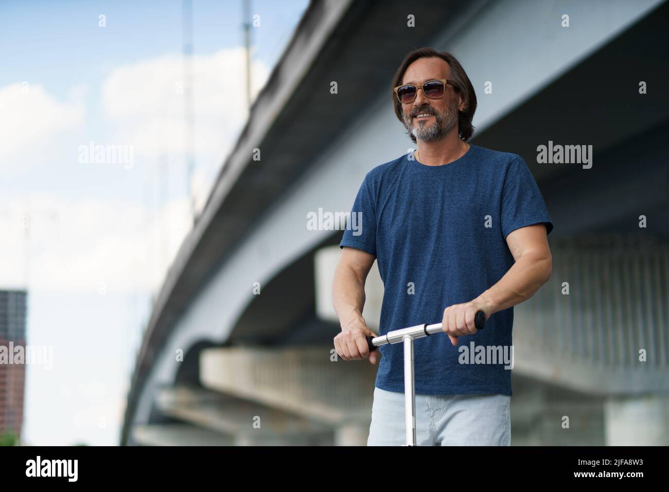 Hübscher, eleganter, reifer Mann mit grauem Bart. Stehen Sie auf einem Stadtroller, und fahren Sie nach der Arbeit im Freien unter der Brücke auf der Straße. Reise-, Lifestyle-Konzept. Stockfoto