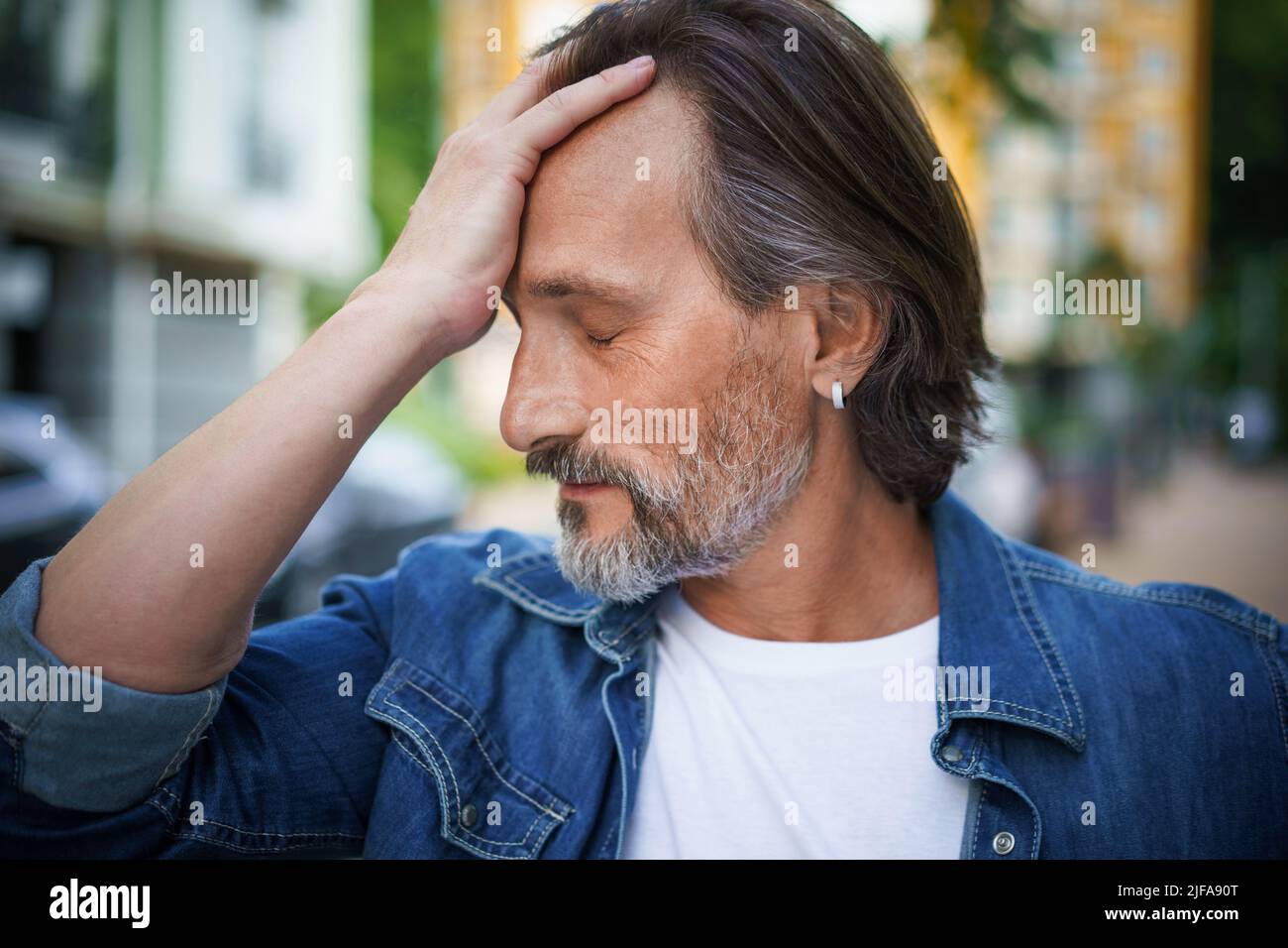 Ein gutaussehender Mann übergeht die Krise des mittleren Alters und berührt seine Stirn, die emotional erschöpft aussieht. Kopfschmerzen, Stress reifer schöner Mann im Freien europäischen alten Stadtstraßen stehen. Stockfoto