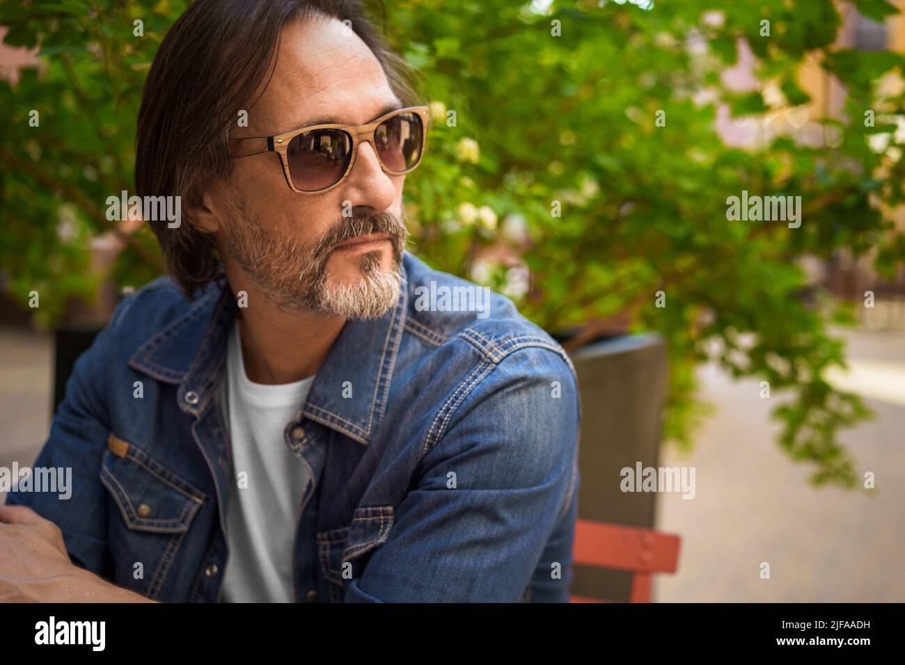 Schöner freiberuflicher Reisender Mann in legerem Denim, der im Stadtcafe im Freien sitzt. Arbeiten im Freien reifer bärtiger Mann mit digitalem Tablet, während er Kaffee genießt. Reisekonzept. Stockfoto