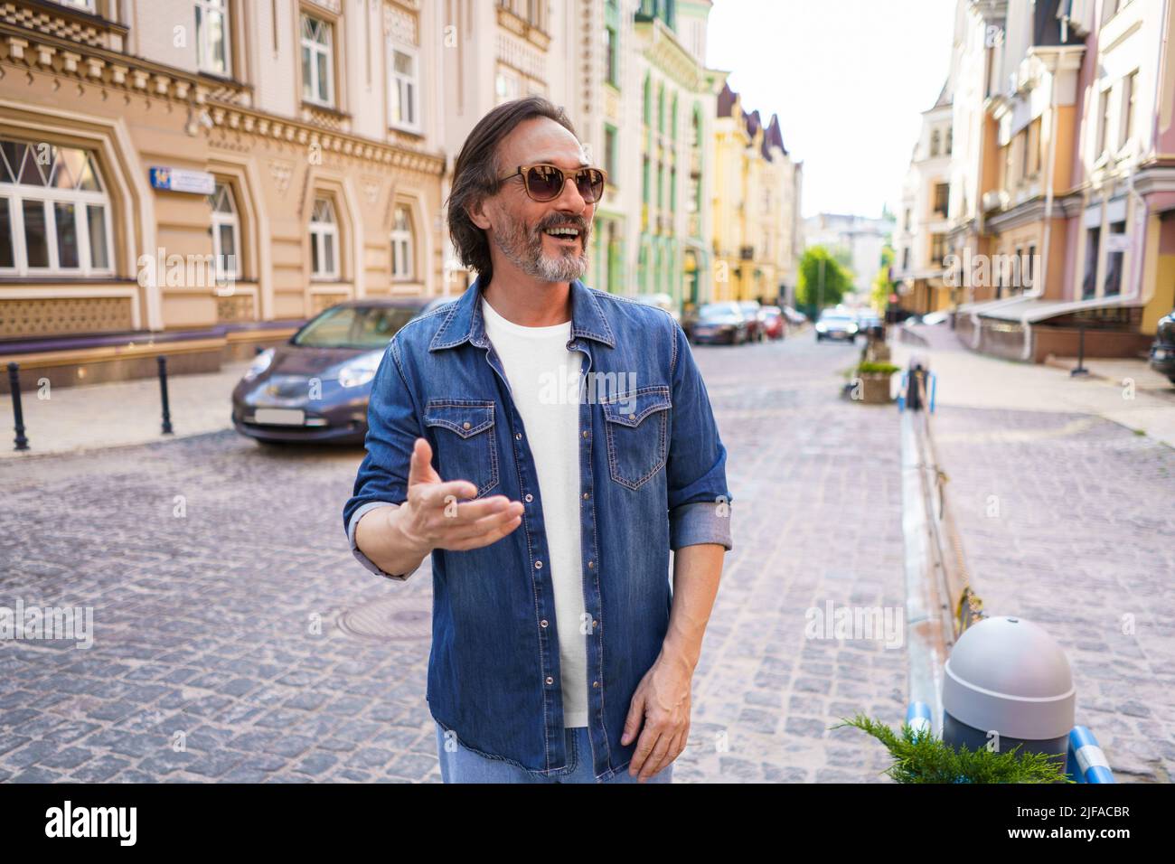 Porträt eines glücklichen, reifen Mannes im Freien in der Altstadt, der lächelnd Jeans-Hemd und Sonnenbrille trägt. Glücklicher, reifer Mann, der an einem Sommertag eine Sonnenbrille trägt und seitwärts nach draußen schaut. Stockfoto