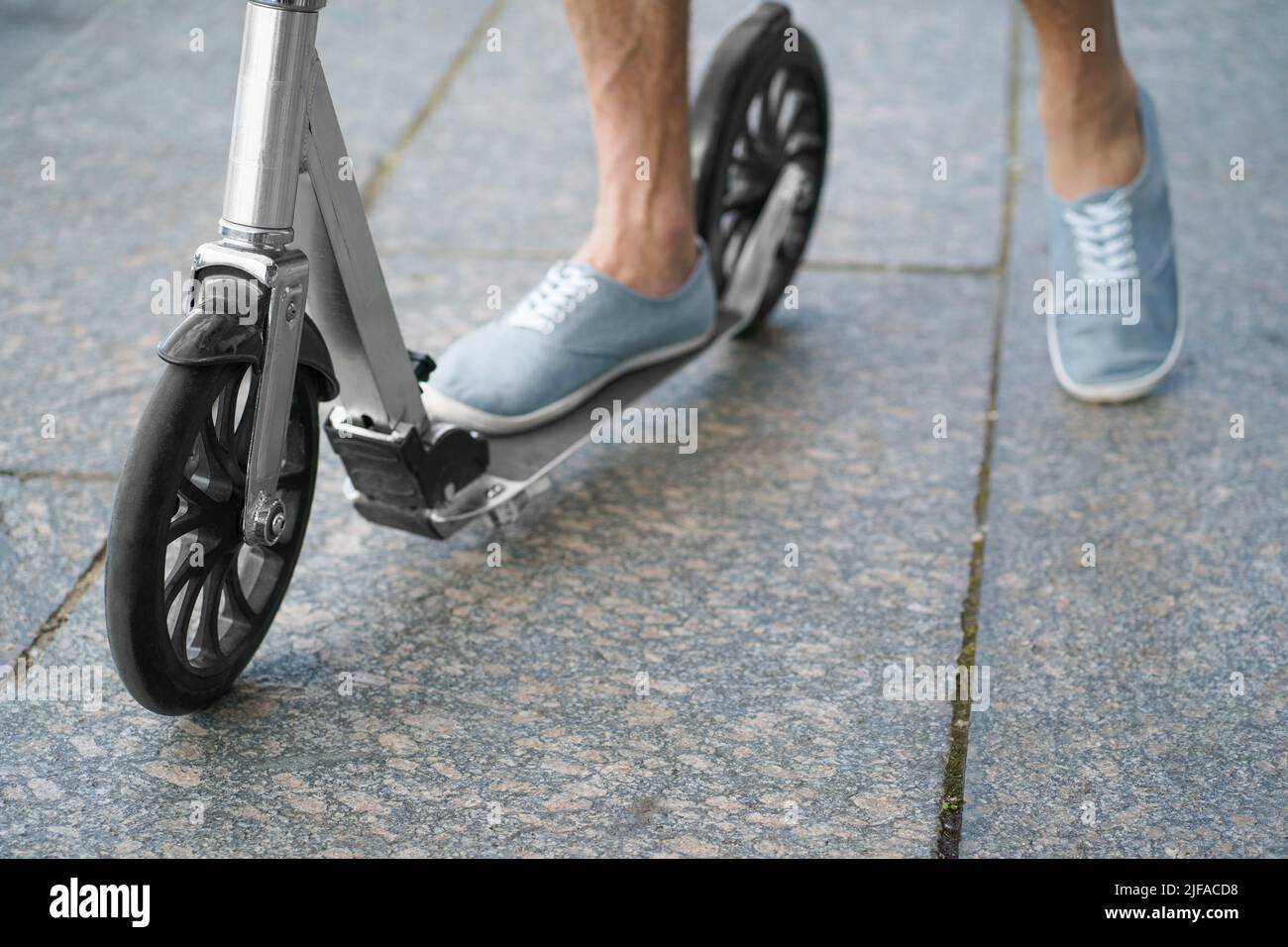 Die Füße eines Mannes in einem stylischen Sneaker stehen auf einem Roller für Erwachsene in der Stadt mit großen Rädern und fahren auf der Straße oder parken nach der Arbeit im Freien. Kein Gesicht sichtbar. Selektiver Fokus auf das Vorderrad. Stockfoto