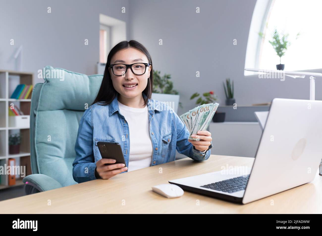 Porträt einer jungen schönen freiberuflichen Frau, die auf die Kamera schaut und lächelt und Telefon und amerikanische Gelddollar hält, online gewinnt. Stockfoto