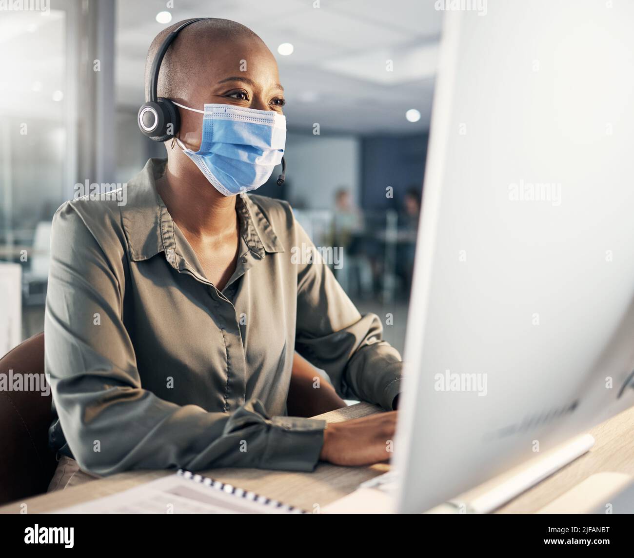 Ein afroamerikanischer Callcenter-Telemarketing-Agent, der Gesichtsmaske als Gesundheits- und Sicherheitsprotokoll trägt und am Headset spricht, während er einen Computer in einem verwendet Stockfoto