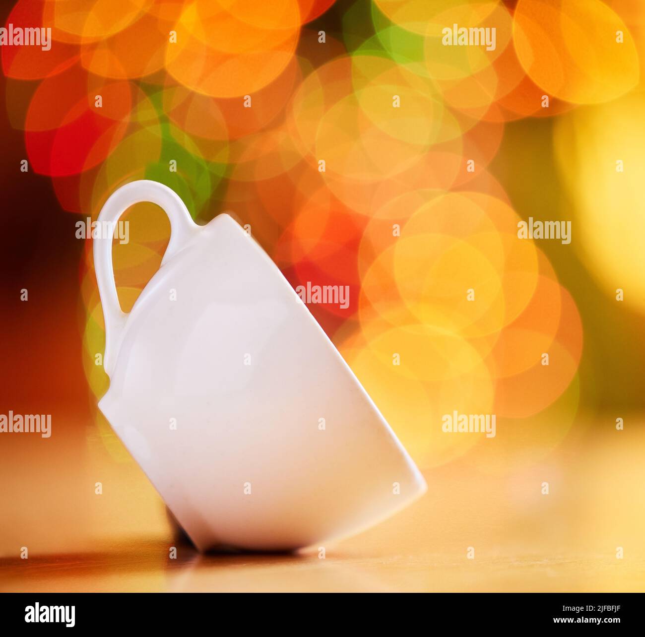 Nahaufnahme einer Tasse isoliert vor einem bunten Bokeh Hintergrund. Weiße Teetasse auf der Seite auf einem Tisch gekippt. Geschirr zum Genießen eines warmen Getränks Stockfoto