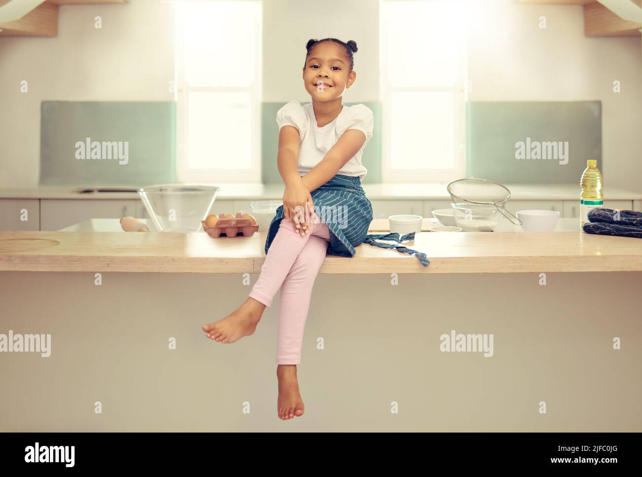 Porträt eines niedlichen jungen Mixed-Race-Mädchens, das mit einer Schürze auf der Küchentheke sitzt und lächelt. Kleine hispanische Mädchen lächelt und sitzt allein Stockfoto