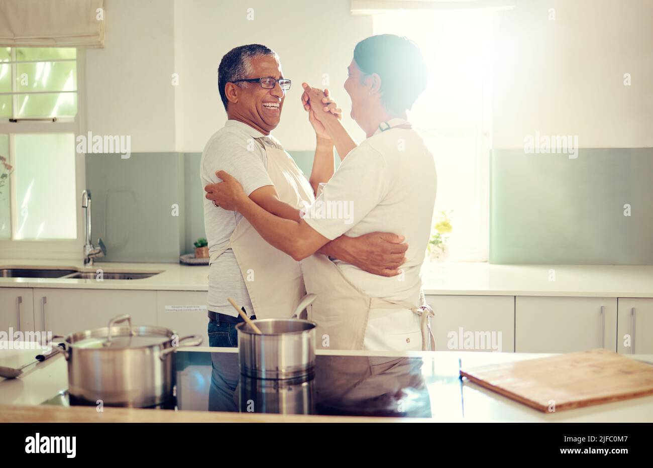 Ein liebevolles Senioren-Mischpärchen trägt Schürzen und hat Spaß beim Stehen und Tanzen in der Küche zu Hause. Romantische reife Mann und Frau lächeln und Stockfoto
