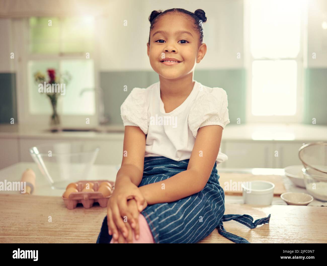 Porträt eines niedlichen jungen Mixed-Race-Mädchens, das lächelnd auf der Küchentheke sitzt und eine Schürze trägt, die nachdenklich aussieht. Kleine unschuldige hispanische Mädchen Stockfoto