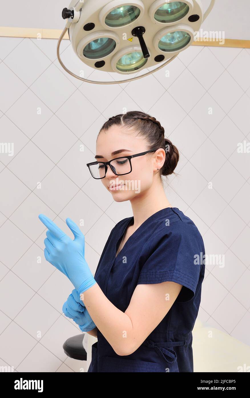 Eine hübsche junge Frau in einem OP-Anzug und einer Brille zieht Gummihandschuhe vor dem Hintergrund einer OP-Lampe an. Stockfoto