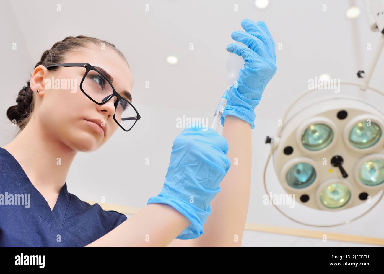 Hübsche junge Frau in einem OP-Anzug und einer Brille mit einer Spritze in den Händen vor dem Hintergrund einer OP-Lampe. Stockfoto