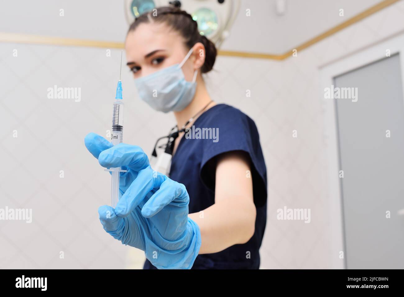 Die hübsche junge Frau in einem OP-Anzug in blauen Gummihandschuhen vor dem Hintergrund einer OP-Lampe hält eine Spritze in den Händen. Stockfoto