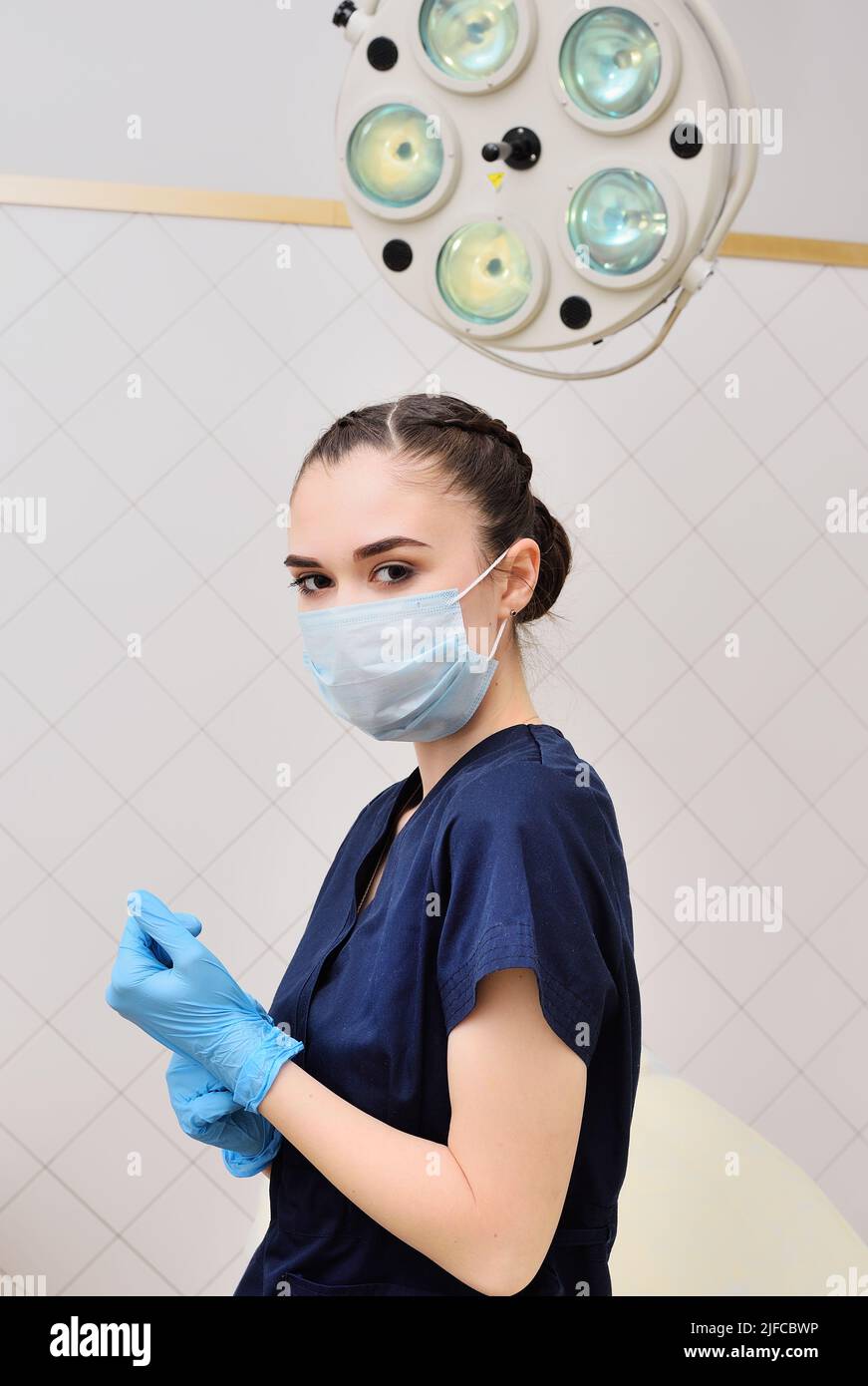 Junge Frau in einem OP-Anzug in einer sterilen Maske vor dem Hintergrund einer OP-Lampe. Stockfoto